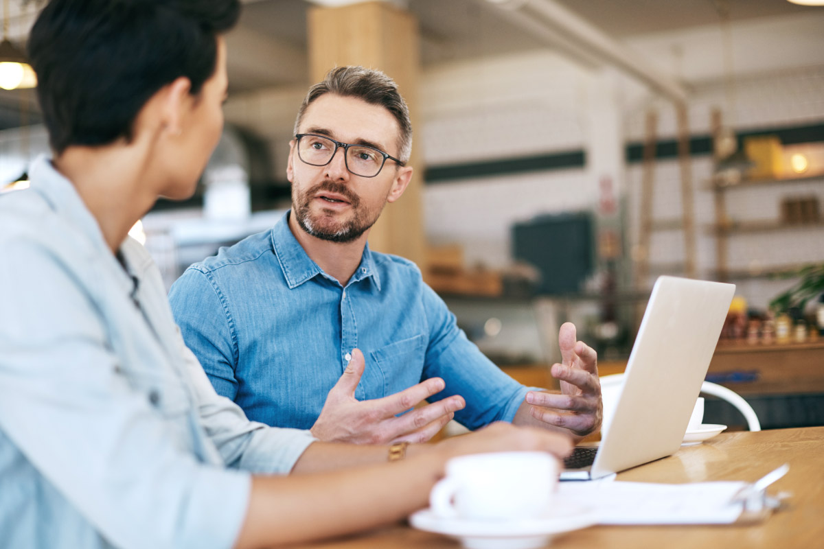 Um homem de cabelos grisalhos com óculos e um laptop na sua frente está conversando com uma mulher sentada em uma mesa de escritório.