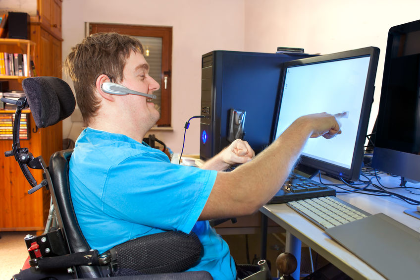 Spastic young man with infantile cerebral palsy caused by complicated childbirth sitting in a multifunctional wheelchair using a computer with wireless headset reaching out to touch the touch screen