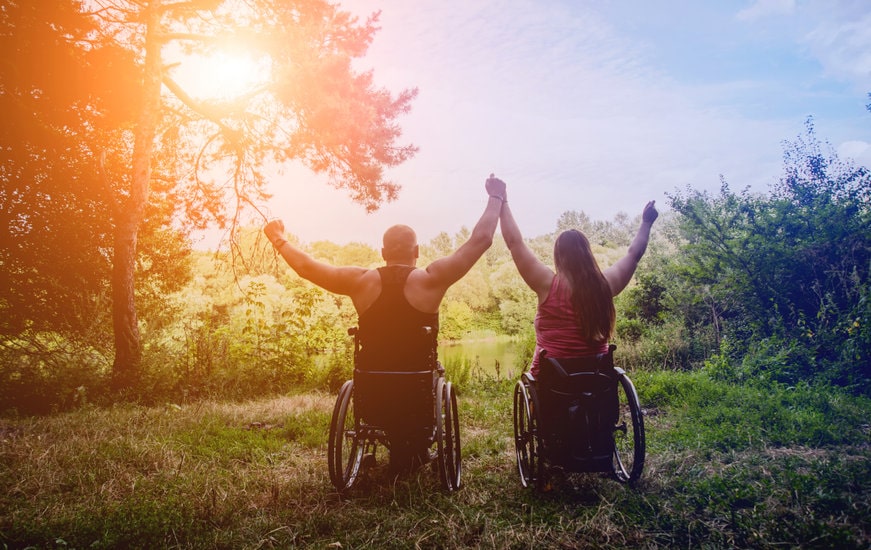 Disabled couple in wheelchairs are holding hands and with arms up in the air enjoying themselves in the forest near the lake.