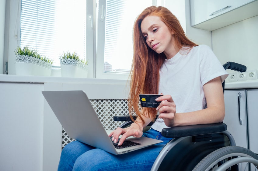 Chica joven pelirroja feliz en silla de ruedas haciendo compras en línea con tarjeta de crédito