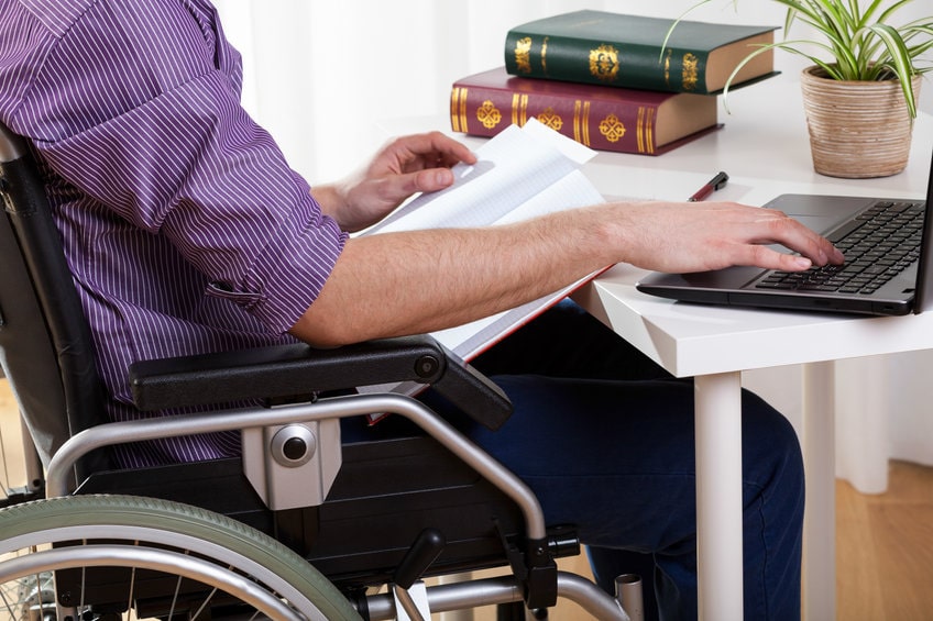 Hombre joven con discapacidad sentado en una silla de ruedas que trabaja con un ordenador portátil en una mesa