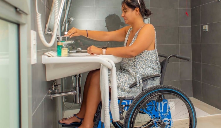 uma mulher com uma deficiência que usa uma cadeira de rodas está usando o lavatório em um banheiro adaptado para pessoas com deficiências.
