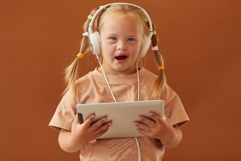 Una niña rubia con síndrome de down sostiene una tableta digital y está mirando a la cámara mientras está de pie contra un fondo marrón