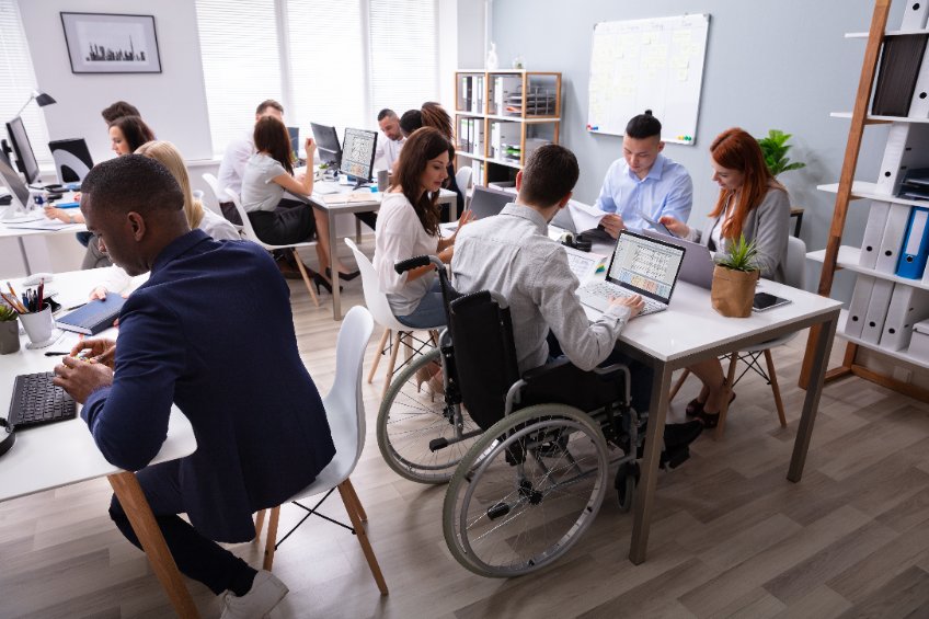 Vista lateral de un empresario con discapacidad sentado en una silla de ruedas con un portátil trabajando en la oficina llena de gente