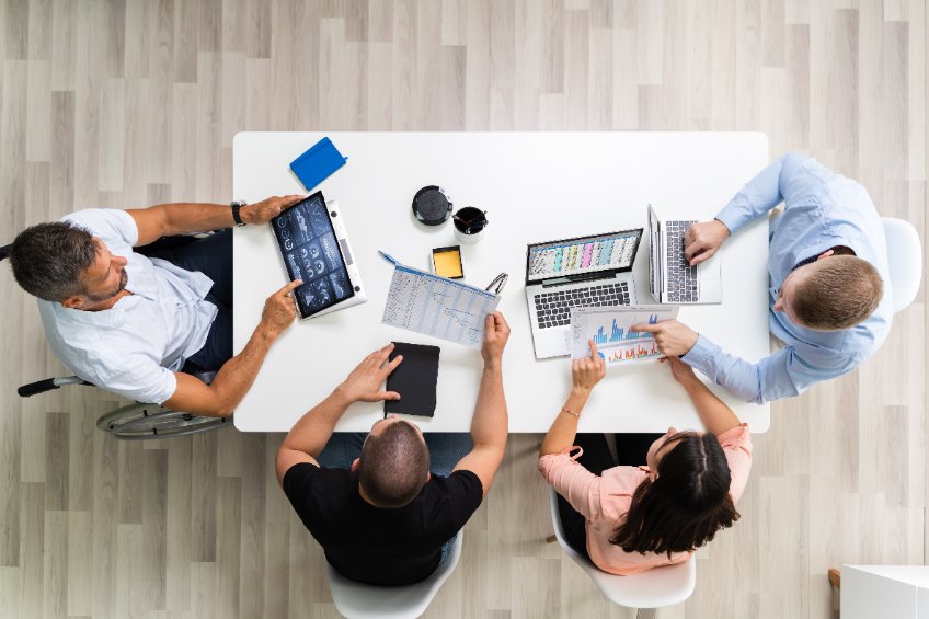 Reunión de negocios informal en una mesa, formada por cuatro personas, una de ellas en silla de ruedas, que utilizan dispositivos electrónicos