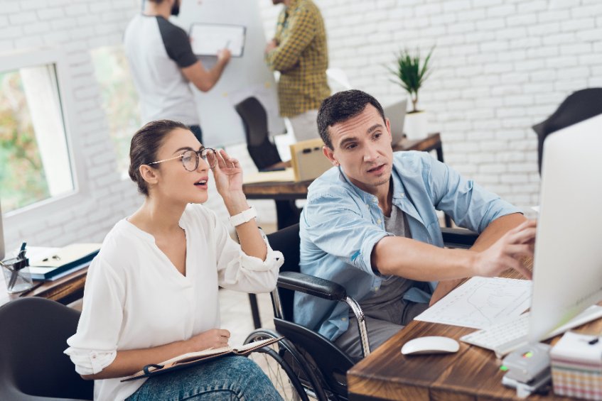 A wheelchair-bound man with a motor disability is showing something on the computer to his co-worker.