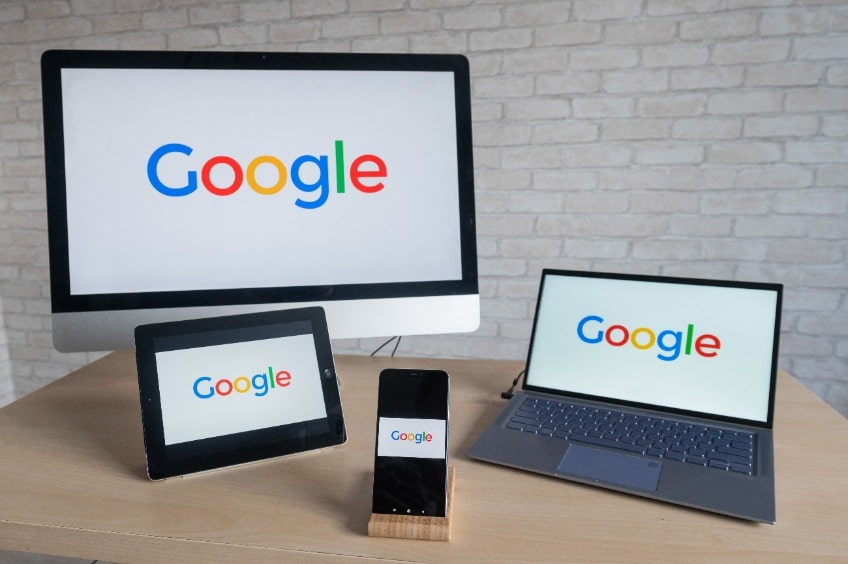 Sobre una mesa de escritorio se encuentran un monitor, un portátil, una tablet y un smartphone, todos ellos mostrando en su pantalla un fondo blanco y el logo de google