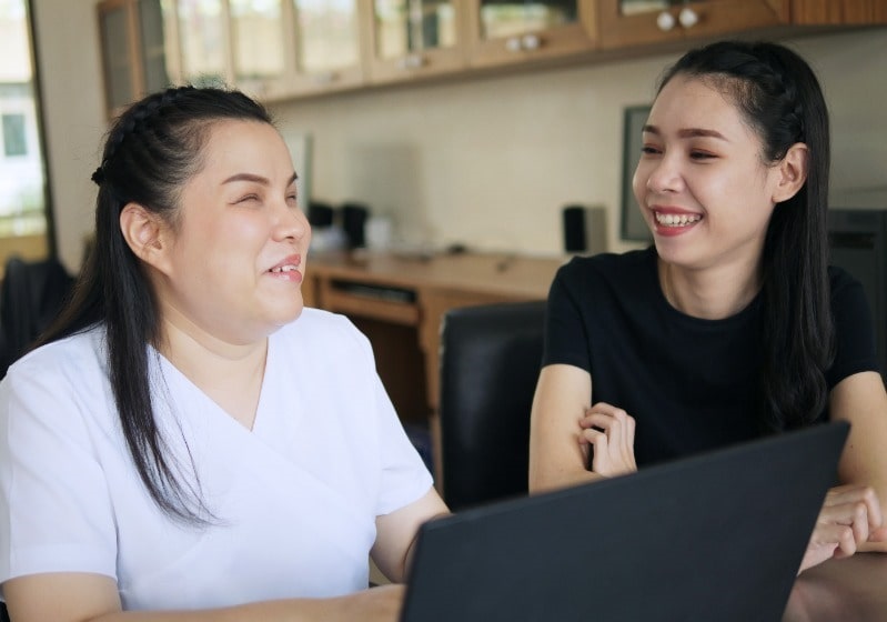 Una mujer asiática con discapacidad visual está interactuando con un ordenador portátil al lado de otra mujer.