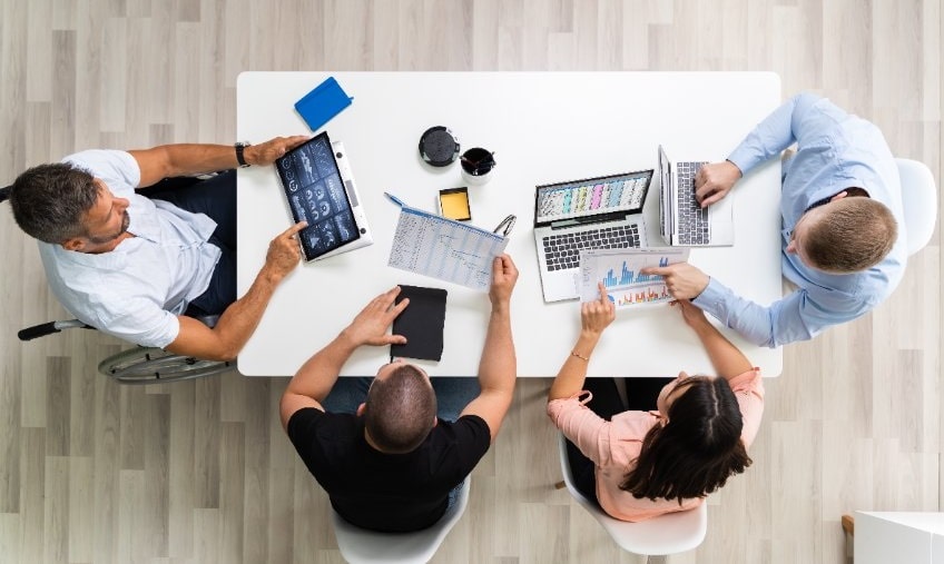 Reunión de negocios informal en una mesa, formada por cuatro personas, una de ellas en silla de ruedas, que utilizan dispositivos electrónicos