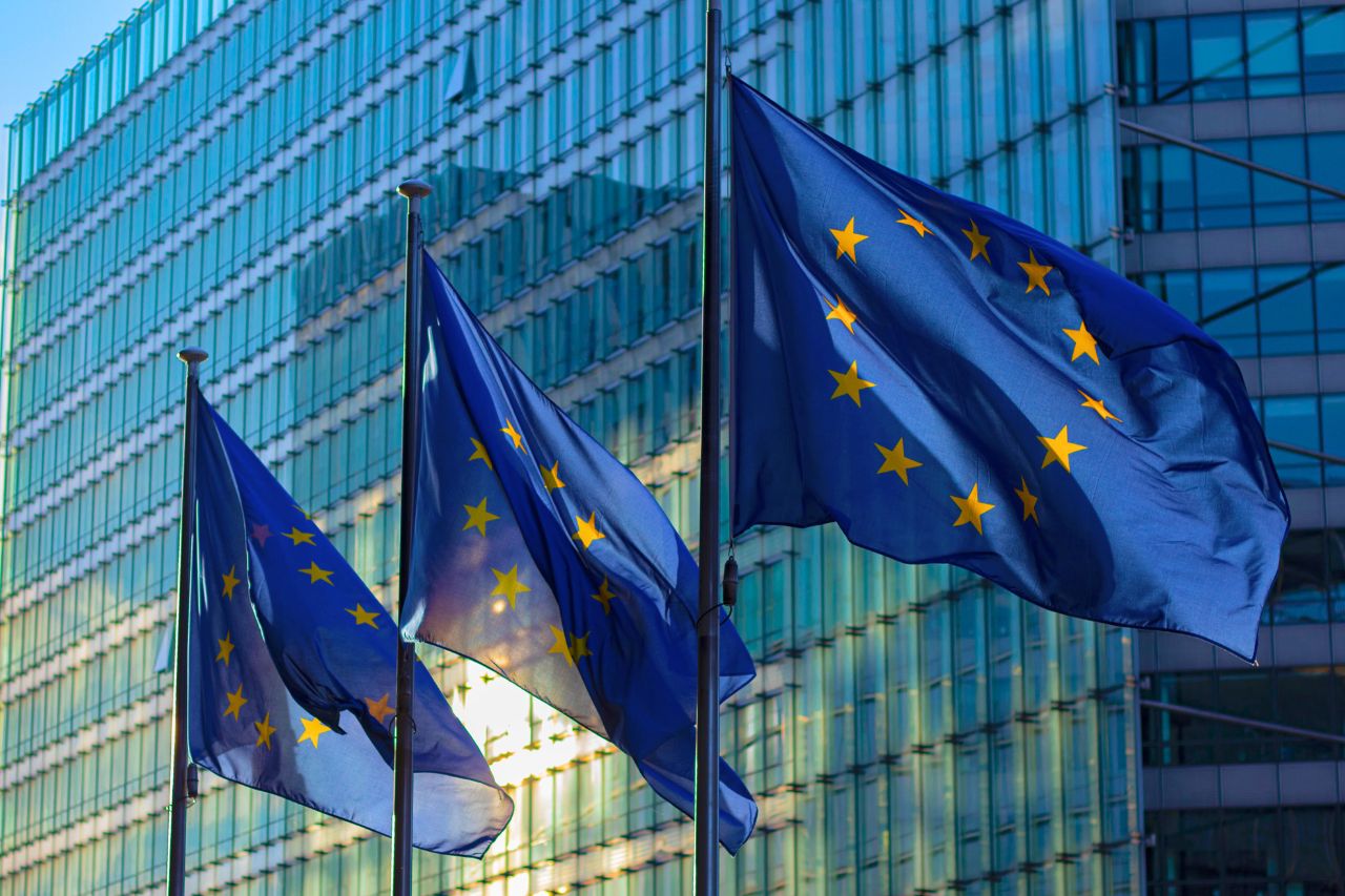 Tres banderas de la Unión Europea ondean frente a un edificio acristalado.