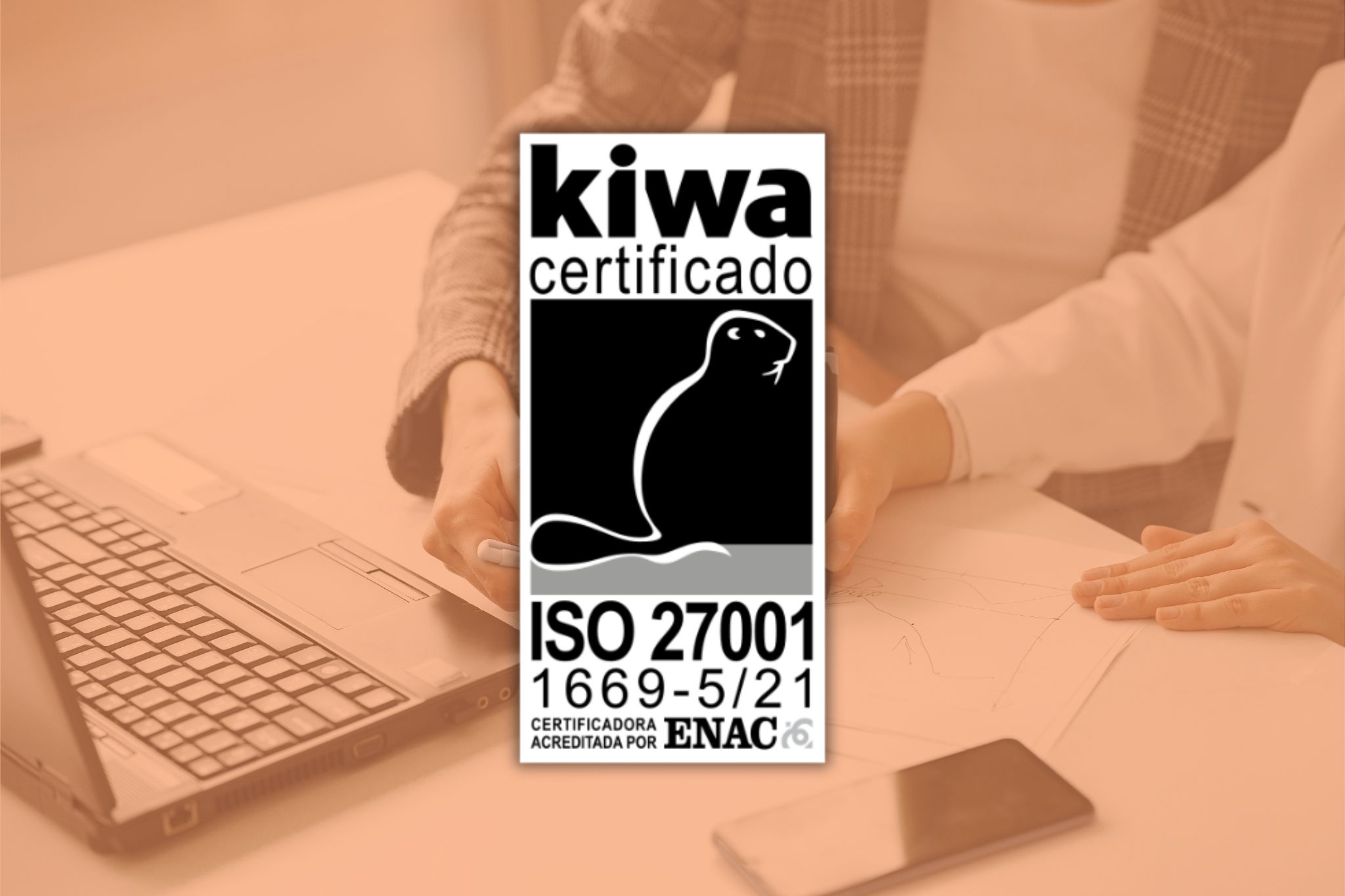 Logo de la certificación ISO 27001 superpuesto sobre una imagen que muestra a dos empleados trabajando en una mesa frente a un ordenador portátil