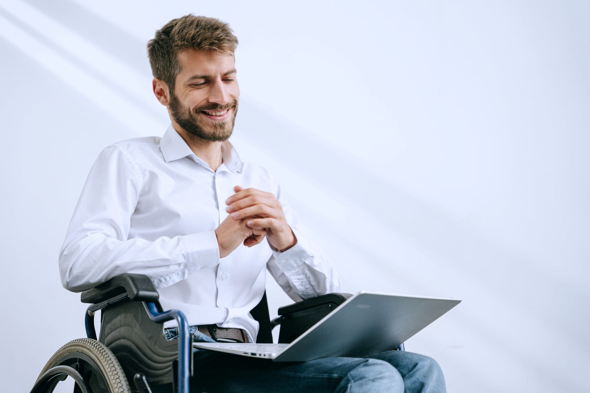 Un hombre joven con discapacidad sentado en una silla de ruedas tiene apoyado un portátil en sus piernas mientras mira su pantalla y sonríe.
