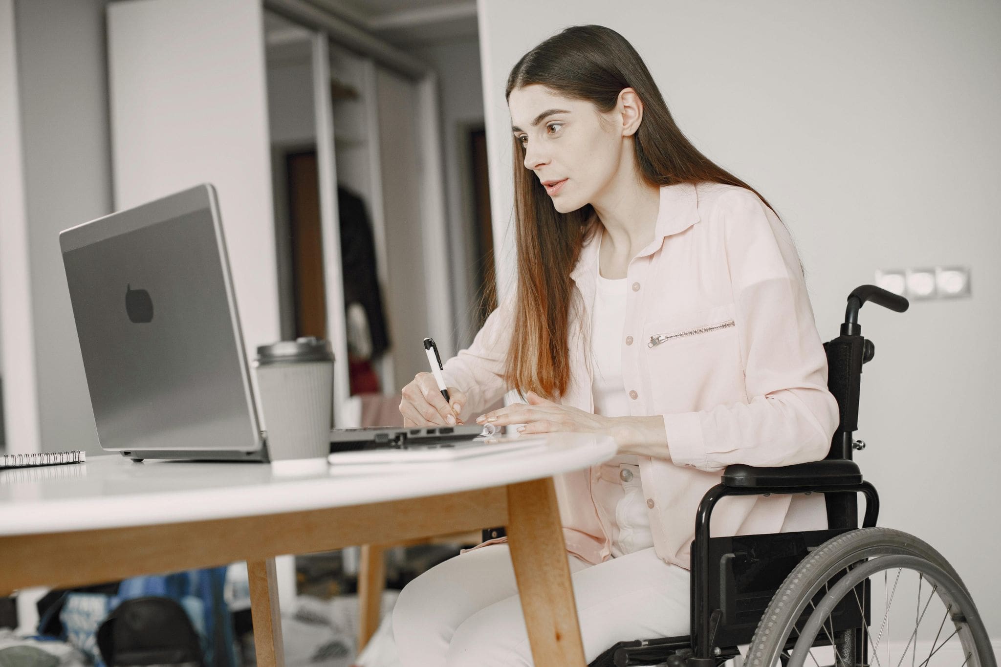 Una mujer con discapacidad está sentada en una silla de ruedas frente a una mesa y su portátil, mientras sujeta un bolígrafo y toma notas en un cuaderno.